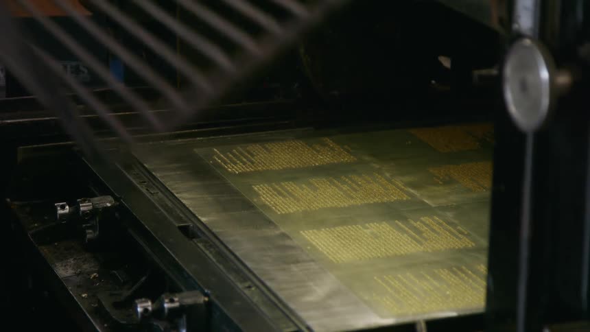 A workers hand quickly pushes a large rectangular metal printing plate covered in gold letters into a printing press then pulls a metal grate over it