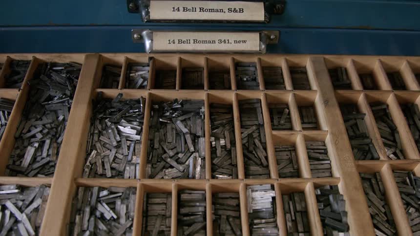 A drawer of an antique printing press cabinet filled with many small silver movable type letters is pulled open