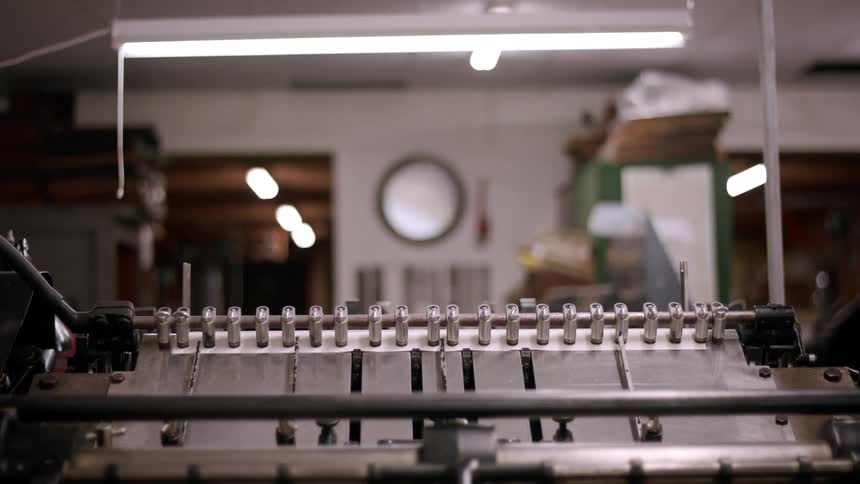 A machine with many metal parts in the foreground feeds and prints on paper which are then pushed out in a line