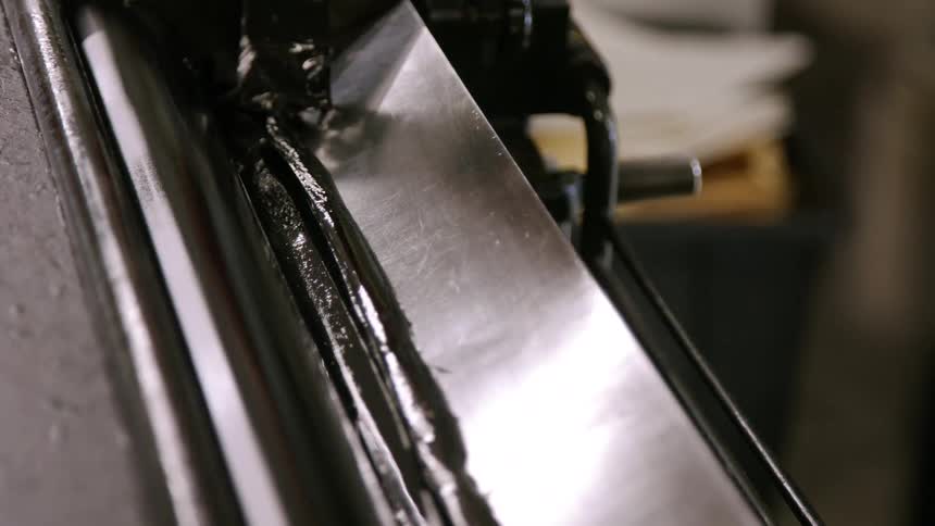 A printing press is shown in a closeup shot with a persons hands pushing a metallic sheet into the machine the sheet is then quickly pulled into the press as the rollers spin