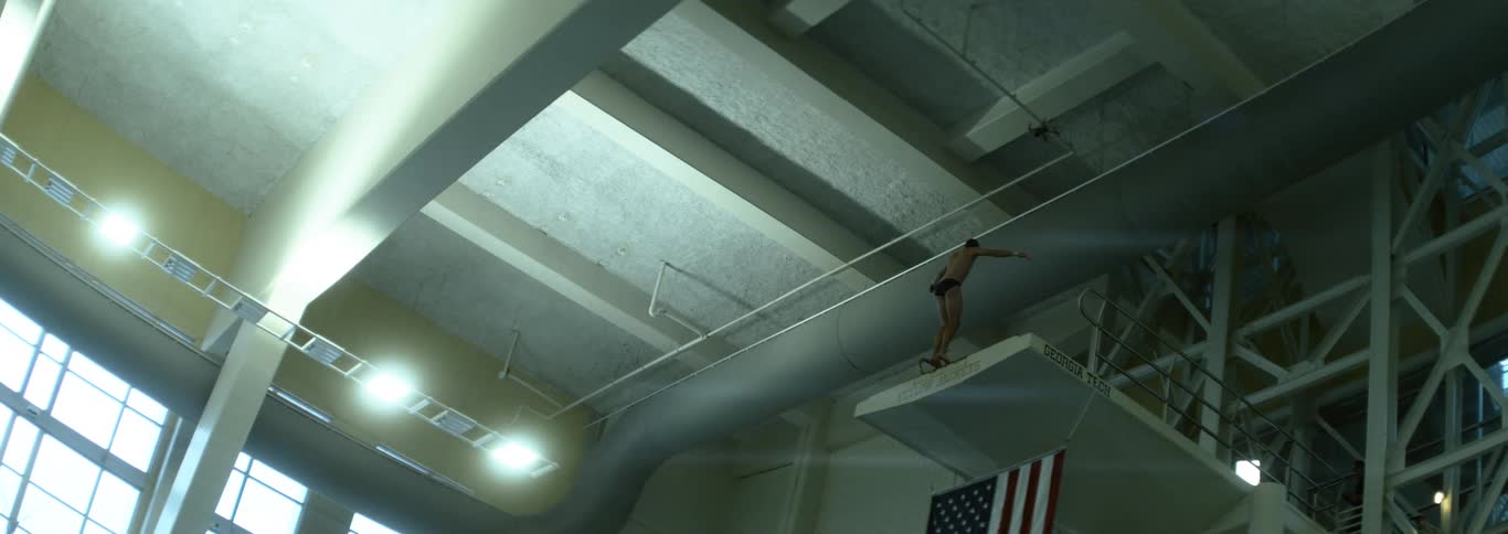 A diver balances on a high platform in a large indoor natatorium