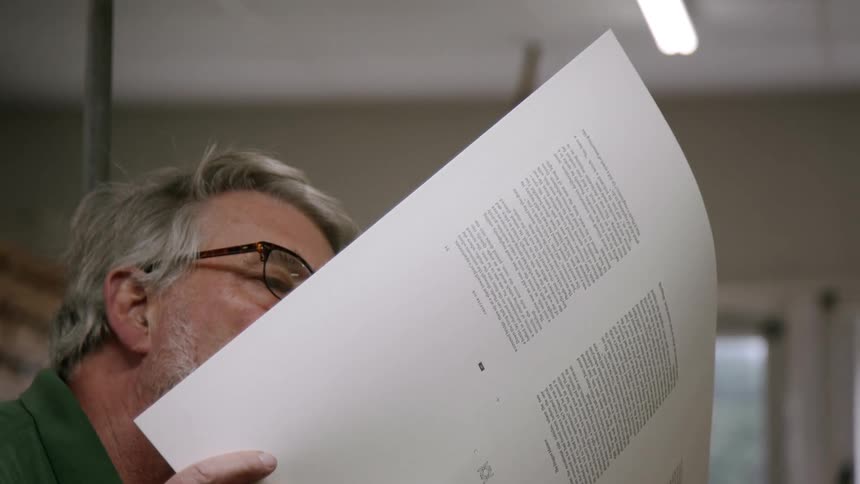 A man with a beard and glasses holds and looks at a large sheet of printed paper