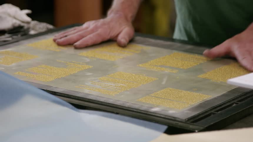 A man is carefully placing a sheet of plastic with printed gold designs onto a larger metal plate he gently presses the film down working to remove any air bubbles