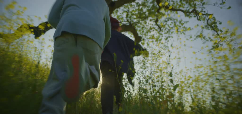 A couple with a dog runs through a field of yellow flowers the man wears a light blue shirt and gray pants while the woman and the dog are in black