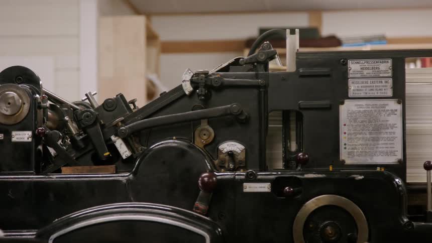 A heavy printing press sits in a workshop its levers and dials catching the light