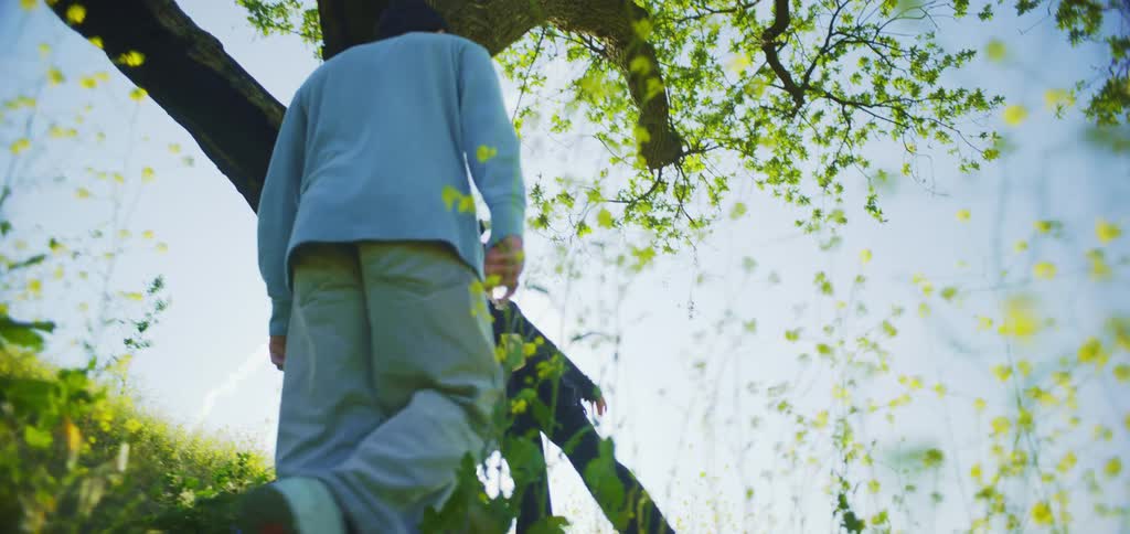 A low angle view shows a man in a light blue sweater walking away from the camera through a field of yellow flowers and green plants as he meets a woman with red hair who touches his face