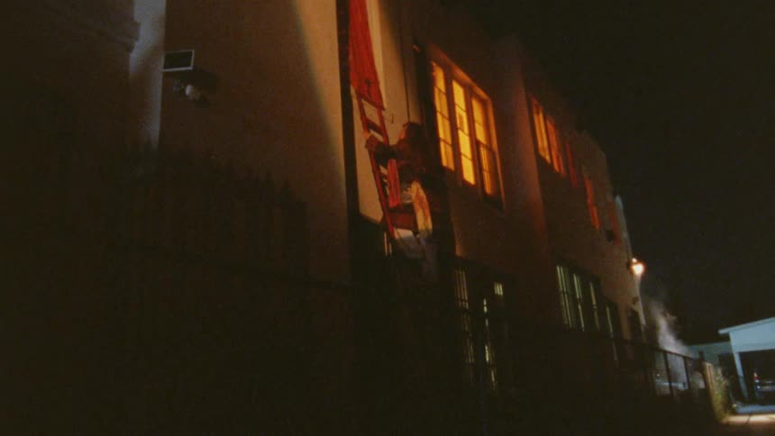 A low angle view at night shows a woman climbing a ladder toward a window on the second story of a dimly lit building