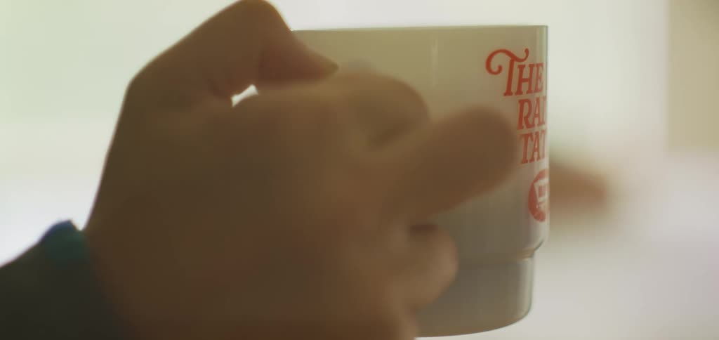 The person drinks coffee while a woman is laying her head on the table