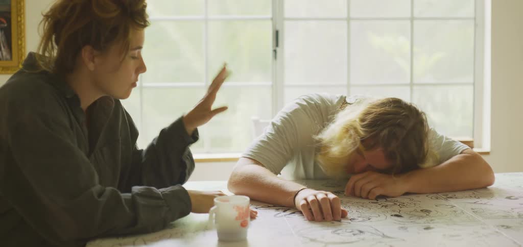A woman offers a white mug to a man with long light hair who is resting his head on a paper covered table as the man then sits up and touches her hand