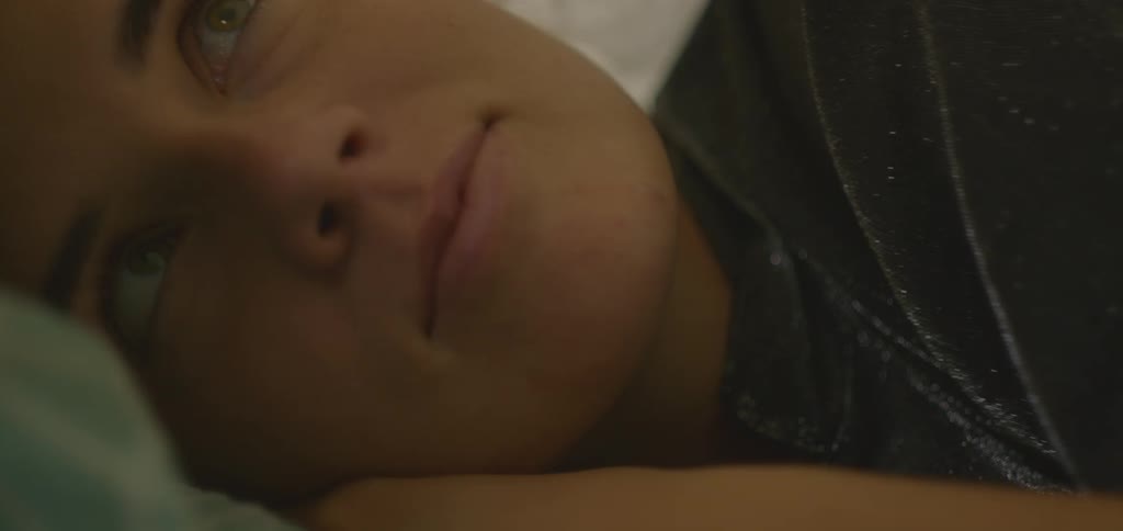 A person is seen from the neck down lying on a bed and looking up at something out of frame the room is dimly lit by a lamp the person is wearing a black leatherlike jacket over a white top