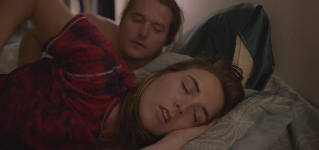 A shot focuses on a person sleeping peacefully in a bed with green sheets and a red checkered comforter the person is curled up in bed tucked under the covers while someone else sleeps nearby