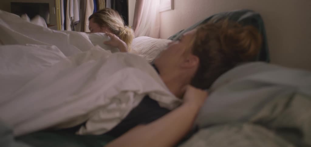 A young woman lying in bed wakes up and turns to wake the woman sleeping next to her who immediately gets up from the bed