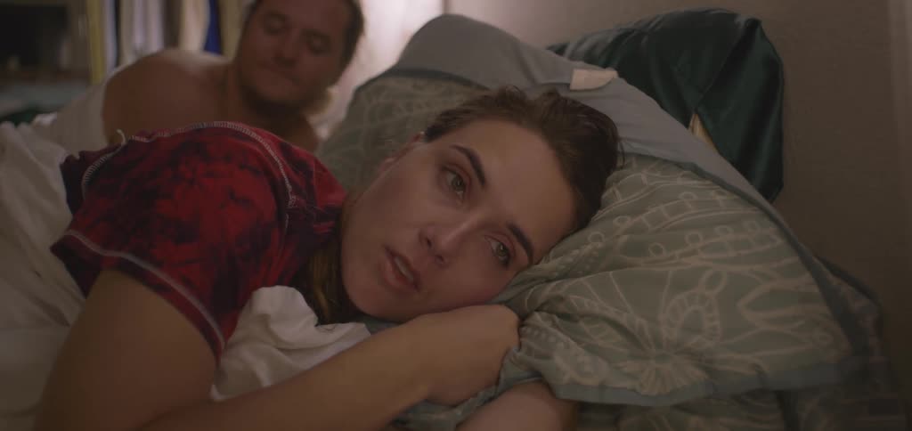 A shot focuses on a person sleeping peacefully in a bed with green sheets and a red checkered comforter the person is curled up in bed tucked under the covers with a pillow tucked under their cheek