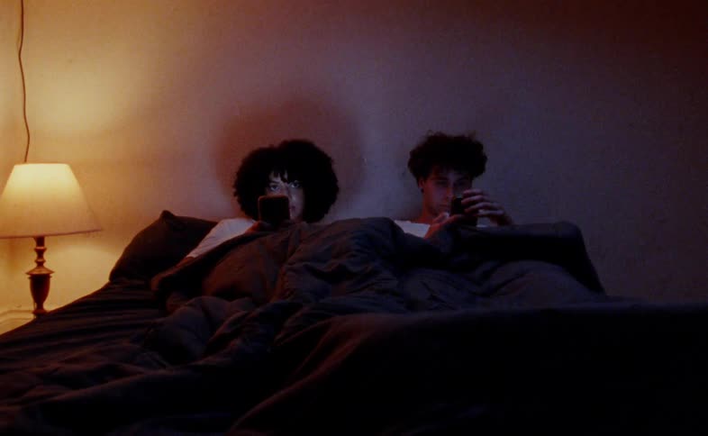 A shot focuses on a couple as they sit in bed together at night illuminated by the glow of their phones they are lying in the dark scrolling through their phones before looking up and smiling at each other
