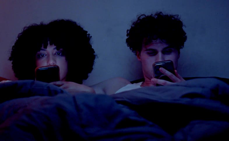 A man and a woman with dark curly hair are lying in a bed under a dark blue comforter with their faces illuminated by the light from their cellphones