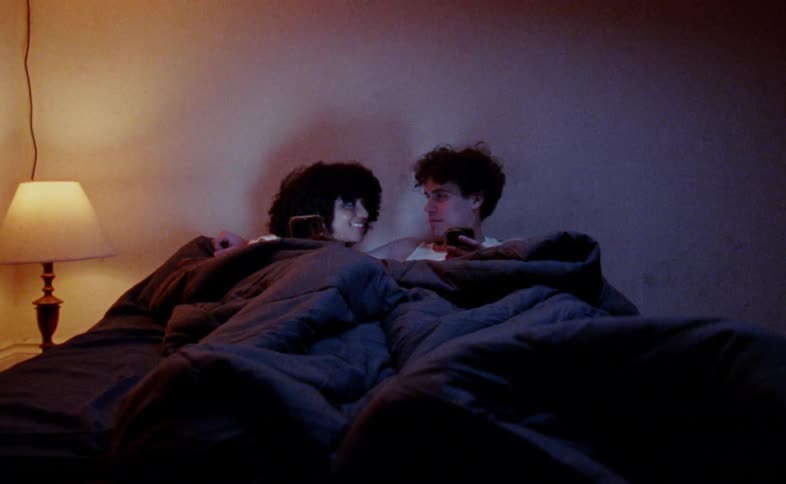 A man and woman are tucked into bed scrolling through their phones in the dark before looking up and smiling at each other the only light in the room comes from a small bedside lamp and the glow of their screens