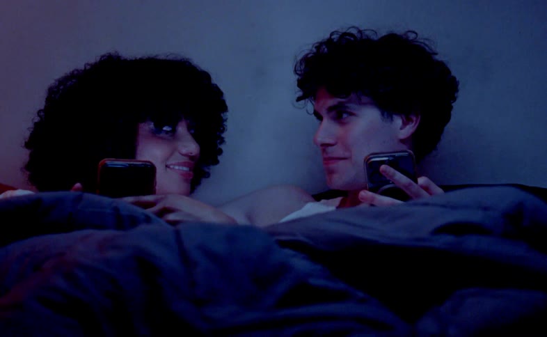 A woman and a man are lying in a bed under a dark blue comforter looking at their cellphones before turning to face each other and getting swallowed up by the blankets