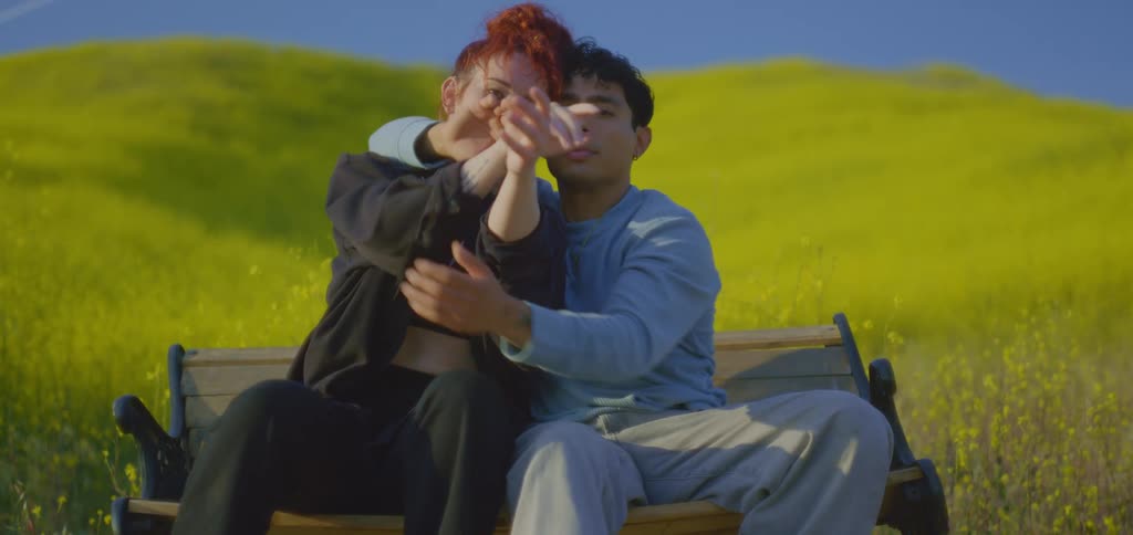 A couple is sitting together on a brown park bench in a wide field of yellow flowers under a blue sky where the man has his arm around the woman and they are moving their hands in a dance