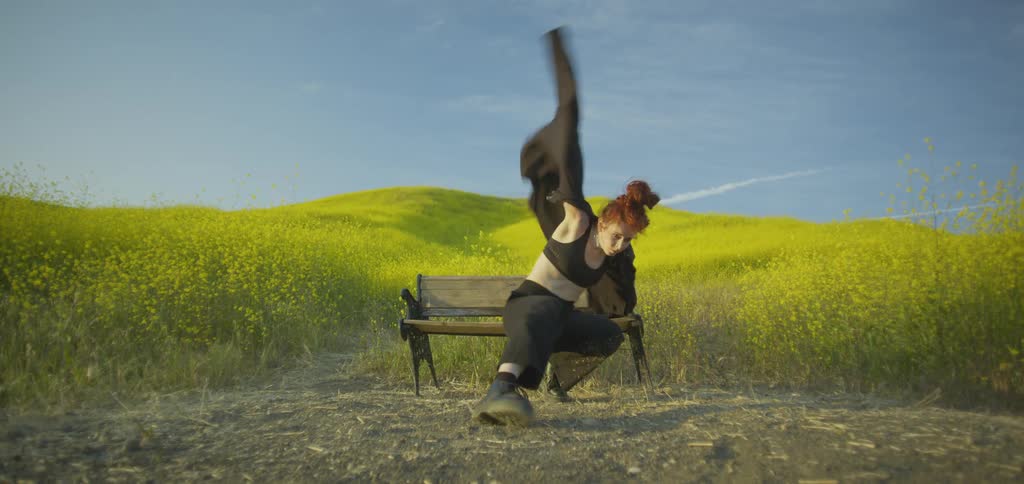 A woman dancing outdoors near a bench in the middle of a tall grassy field