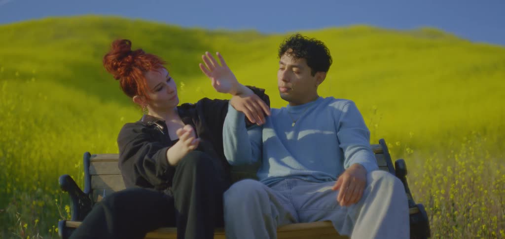 A young man and woman perform an expressive interpretive dance while sitting on a bench in a field of yellow wildflowers under a clear blue sky