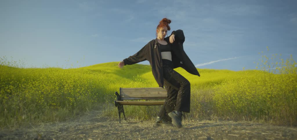 A woman with her red hair in a bun is dancing and stretching with a fluid expressive style next to a wooden bench in a wide field of yellow flowers under a blue sky
