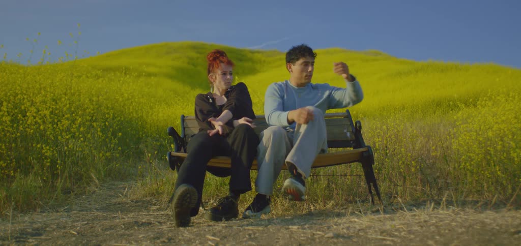 Two people sit on a bench in a field of yellow plants and move their body while facing the camera