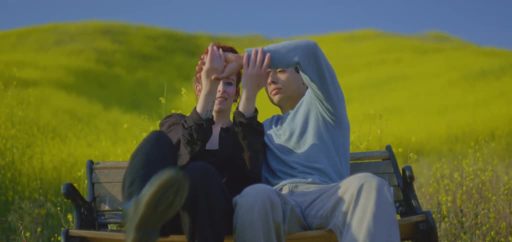 A couple is sitting on a brown park bench in the middle of a yellow field as the woman rests her head on the mans shoulder and they playfully reach their hands up and around