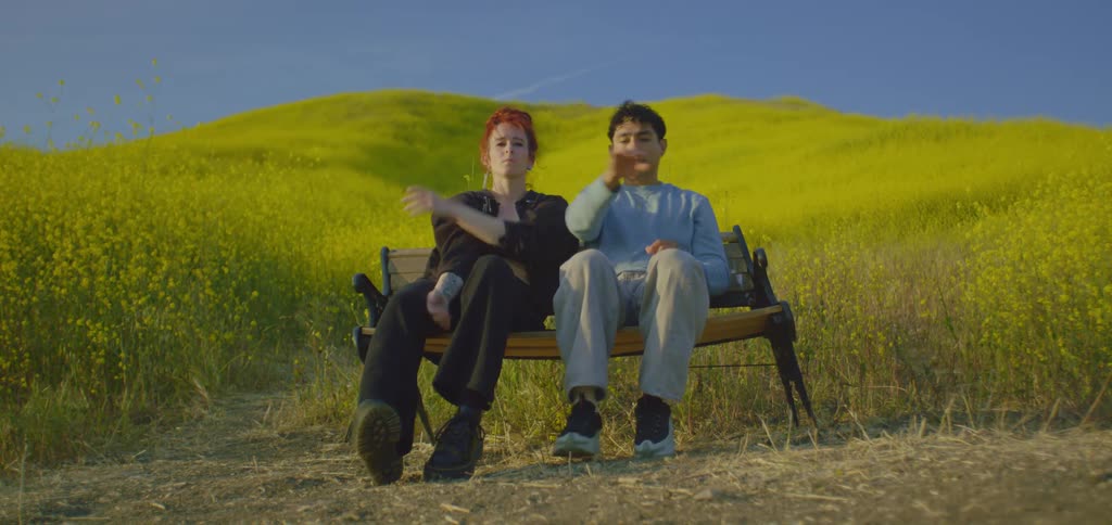 A young boy and a young girl with red hair sit on a bench in a grassland at midday each showing different hand dance moves