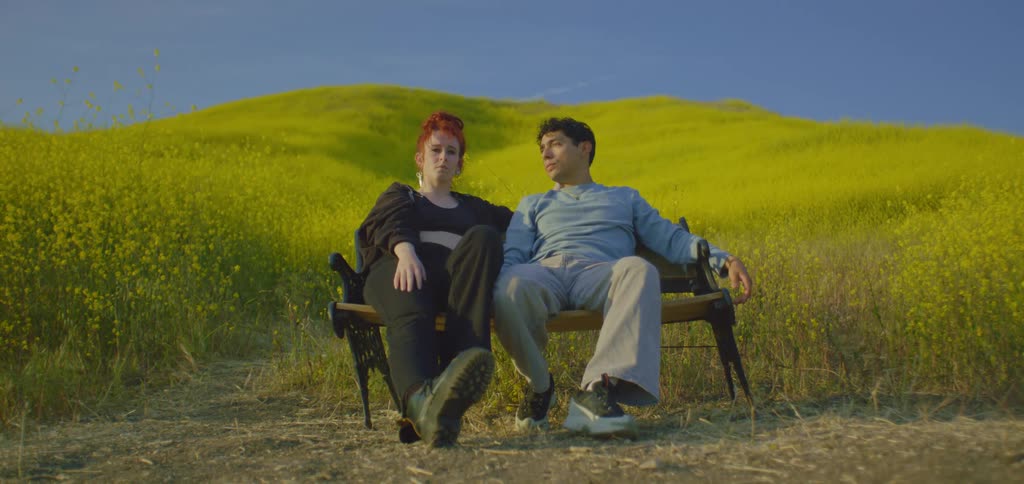 A shot focuses on a couple as they sit on a bench in a field of yellow flowers the woman is raising one hand in a joyful gesture with the man sitting calmly beside her