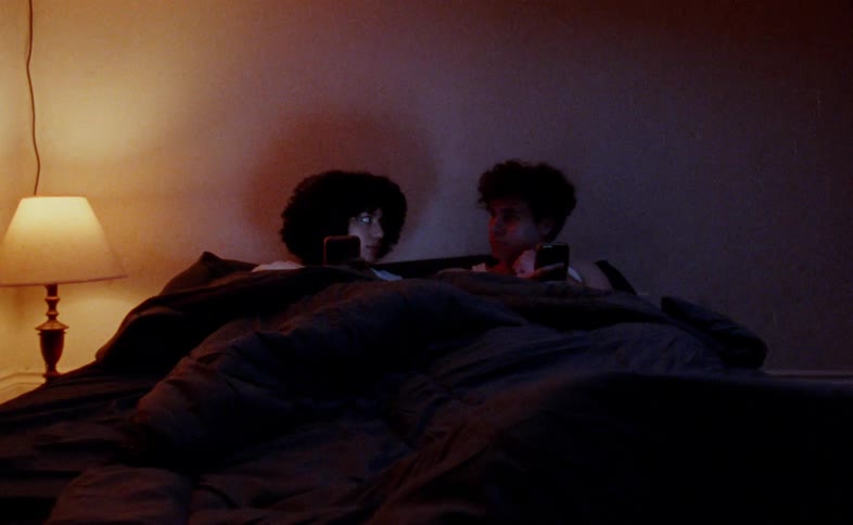 A couple a man and a woman lying in bed under a blanket using their phones with the man saying something