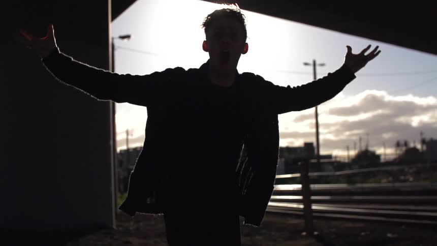 Sun glaring in the background as a young man spreads out his arms and leans towards the camera under an overpass