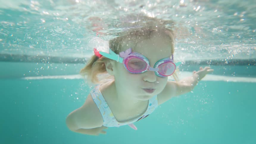 Little girl in pink and teal goggles swimming underwater in a swimming pool