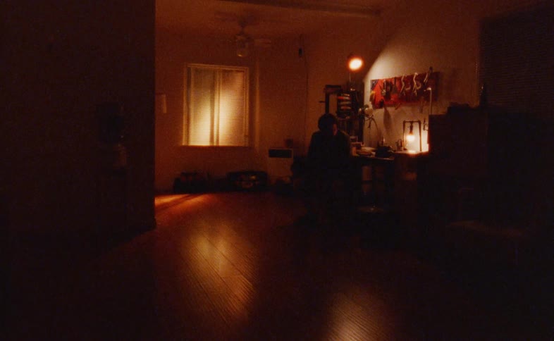 The video shows a wide shot of a dimly lit room with a person sitting at a desk on the right side of the frame the main source of light is a lamp on the desk and sunlight streaming through a window with closed blinds