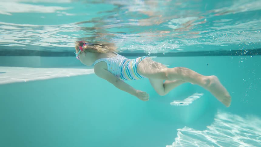 A young girl wearing goggles swims in a pool on a sunny day