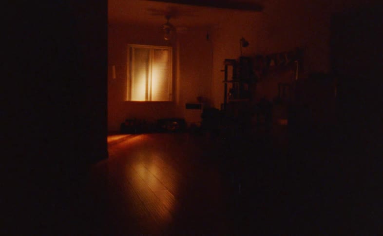 A large silhouetted person walks from the left to the right across a dark wooden floor passing in front of a brightly lit window that reflects onto the ground
