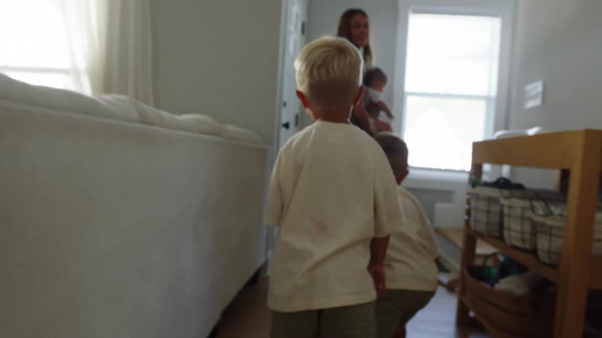 A mother carrying a baby is seen from a low angle as she runs away from the camera and is followed by two young children who are running after her