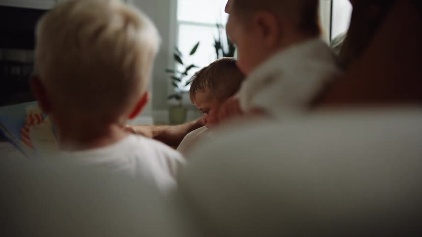 A father sits on a couch at midday carrying one of his toddler sons on his lap while the mother holds an infant and another son sits beside her as the father reads them a storybook