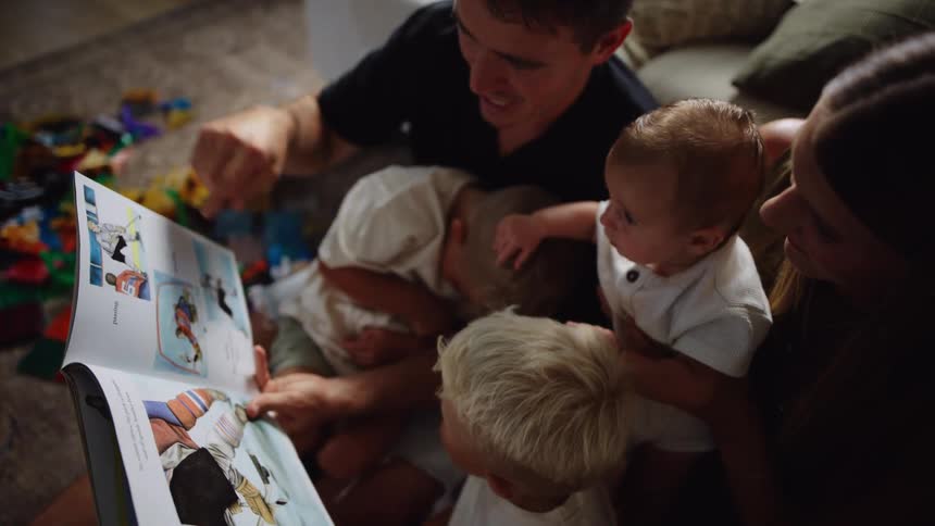 Young family sits on the floor in their living room while the father reads a story out loud