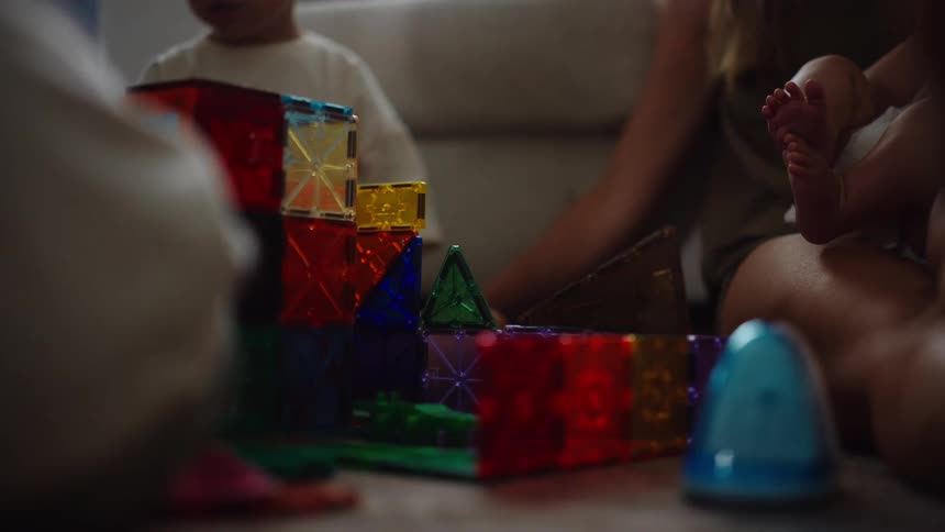 The video shows a family including a young child and an infant playing together they are on a carpeted floor building a structure with colorful magnetic tiles the video has a soft warm light and a familyoriented feel