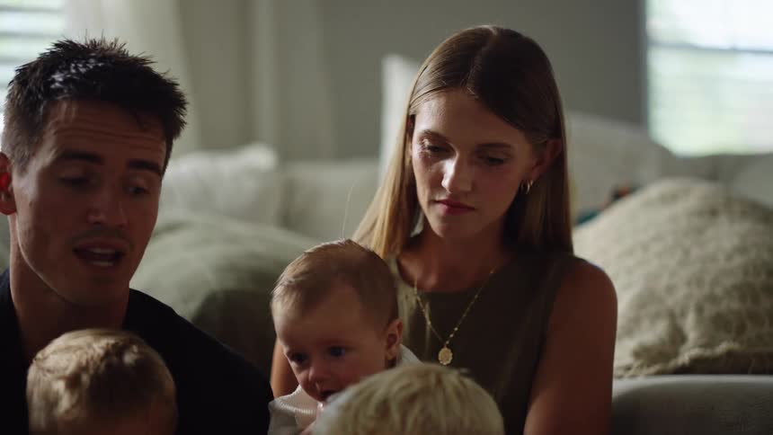 The video shows a man and a woman sitting on a couch with a baby on each of their laps they are looking at the babies and appear to be smiling and interacting with them