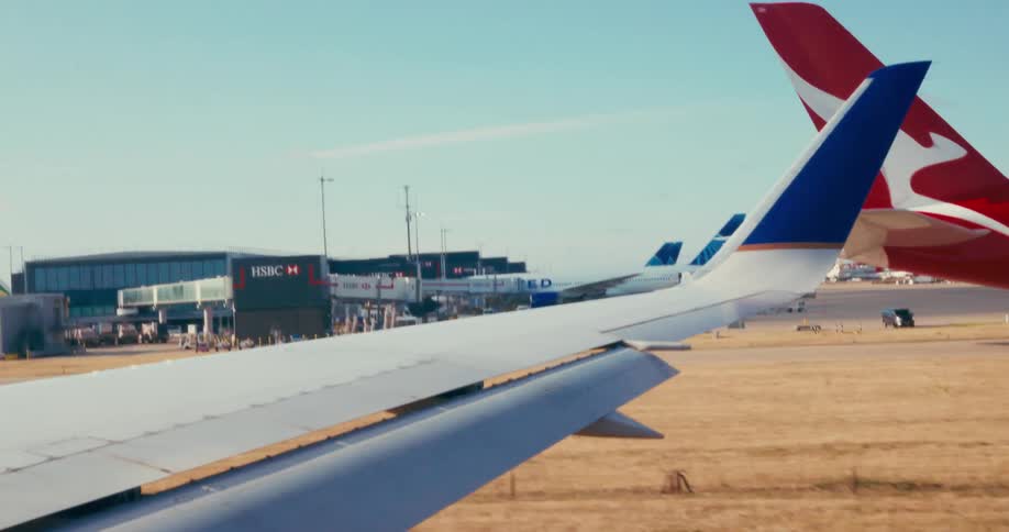 A shot from a moving car shows a planes wing on the left and the tails of two other planes on the right the planes are parked on the tarmac with airport terminals in the background