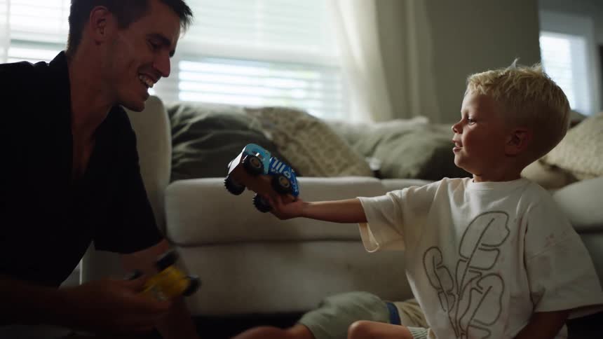 A father is on the floor playing with his two young sons the older son who has light hair playfully pushes a toy car toward his dad