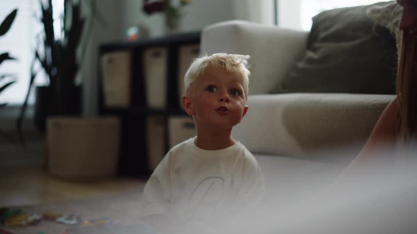 A blondehaired child is sitting on the floor in a living room playing with toys lightcolored fog is seen rolling across the floor possibly coming from a fog machine
