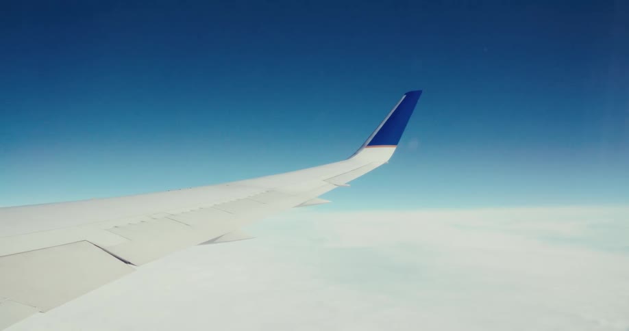 A high angle view from a plane shows a wing with a blue and white tail fin as the aircraft flies above a thick layer of white clouds under a clear blue sky