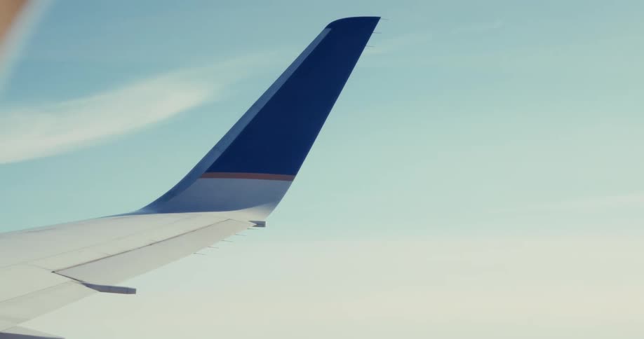 View from a passenger window shows a jet wing slicing through pale blue sky and soft cloud bands