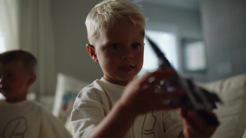 A child with blond hair holds a dark toy airplane looking at the camera and then turning his head to look at a second child playing behind him