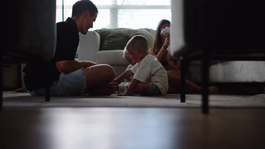 A low angle view shows two parents and their two young children playing together on the floor with toys in a brightly lit room with large windows and a sofa