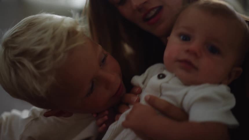 A mother lovingly interacts with her two children in a sunlit room she holds a baby in her arms while a young boy with light hair leans in to kiss the babys cheek