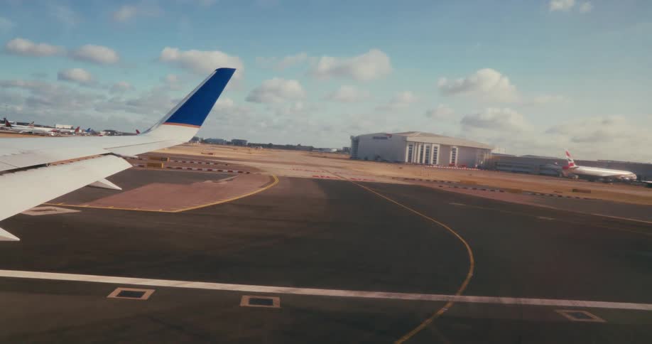 A large airplane wing with a blue tail fin is visible in the foreground as the aircraft moves along a runway at a busy airport with several hangars and other planes in the background