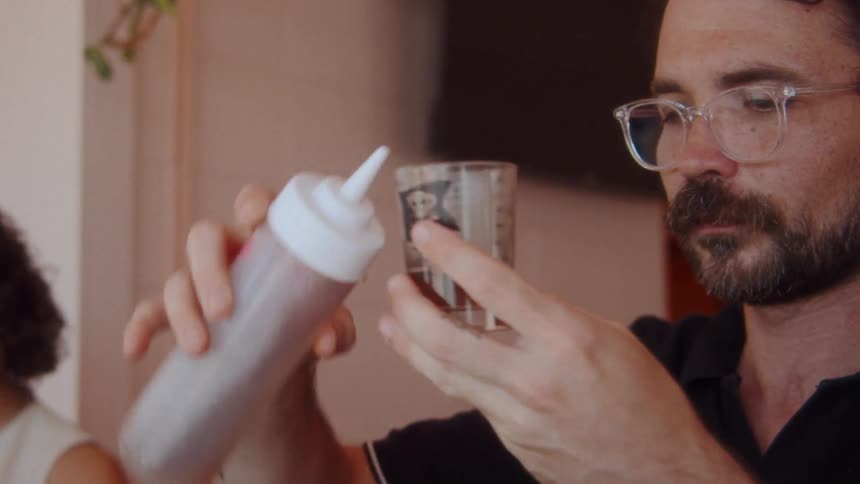 A man in glasses pours a liquid from a squeeze bottle into a glass